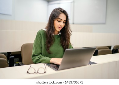 Portrait Of A Beautiful, Young And Intelligent-looking Indian Asian Woman Student Wearing A White Shirt And Green Tracker Smiling As She Works On Her Laptop In A University Classroom. 