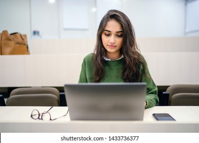 Portrait Of A Beautiful, Young And Intelligent-looking Indian Asian Woman Student Wearing A White Shirt And Green Tracker Smiling As She Works On Her Laptop In A University Classroom. 