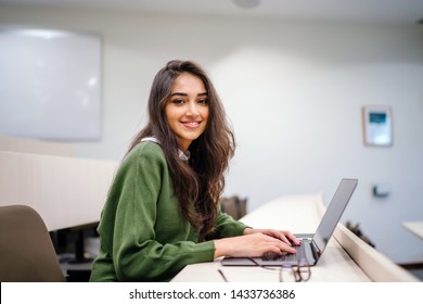 Портрет красивой, молодой и умно выглядящей индийской азиатской студентки в белой рубашке и зеленом трекере, улыбающейся, когда она работает на своем ноутбуке в университетском классе. 