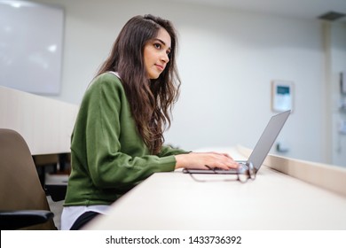 Portrait Of A Beautiful, Young And Intelligent-looking Indian Asian Woman Student Wearing A White Shirt And Green Tracker Smiling As She Works On Her Laptop In A University Classroom. 