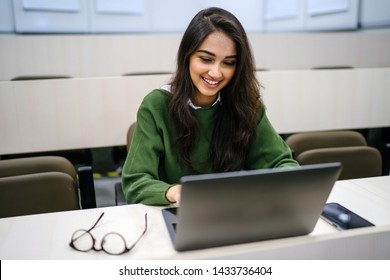 Portrait Of A Beautiful, Young And Intelligent-looking Indian Asian Woman Student Wearing A White Shirt And Green Tracker Smiling As She Works On Her Laptop In A University Classroom. 