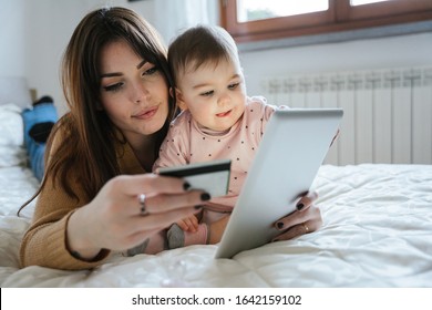 Portrait of a beautiful young mom with a baby girl lying on the bed in a moment of intimacy while buying products online with the tablet and holding her credit card in hand - Millennial with daughter - Powered by Shutterstock