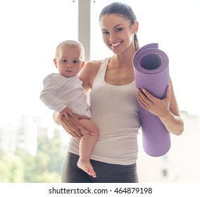 Portrait of beautiful young mom in sports wear holding her charming little baby and yoga mat, looking at camera and smiling while standing against window