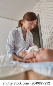 Portrait Of Beautiful Young Mother Changing Clothes On Her Baby In Light Bedroom. Lovely Mum Changing Baby's Diaper.