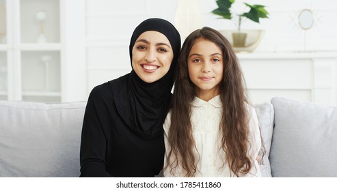 Portrait Of Beautiful Young Muslim Mother In Headscarf Smiling To Camera While Sitting With Cute Teen Daughter On Sofa In Living Room. Pretty Arabian Woman In Hijab And Small Teenage Girl On Couch.