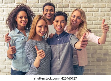 Portrait of beautiful young people of different nationalities showing Ok signs, looking at camera and smiling, against white brick wall
