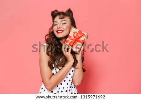 Portrait of a beautiful young pin-up girl wearing dress standing isolated over pink background, showing gift box