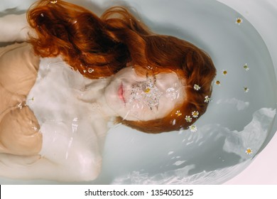 Portrait Of A Beautiful Young Red-haired Taking Bath In Spa Centre, Keeping Face Under The Water. Sexy Young Lady With Bare Shoulders And Wet Coppery Hair