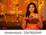 Portrait of a beautiful young smiling Indian woman in traditional dress holding plate full of oil lamp light or diya with festive lights decoration in the background celebrating diwali.