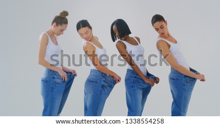 Portrait of beautiful young smiling women of different ethnicities with perfect firm and slim body in white tank shirts showing weight loss by wearing old jeans isolated on white background.
