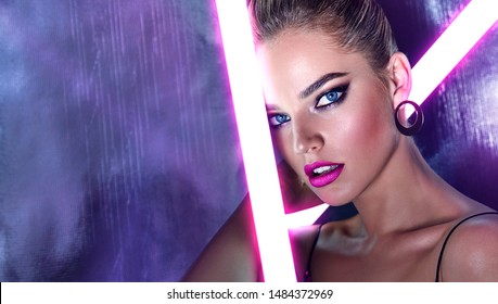 Portrait of a beautiful young tanned girl in big sunglasses in a pink shiny dress in the studio on a silver background with a pink and purple hue. Glowing fluorescent lamps in the face.