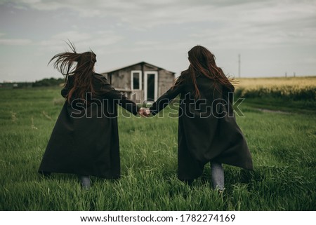 Portrait of beautiful young two women sisters walking and holding hands in black coats and jeans with long hair in a green field in summer. Advertising of stylish clothes. Romantic, family love.