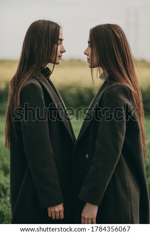 Portrait of beautiful young two women sisters in black coats and jeans with long hair in a green field in summer. Advertising of stylish clothes.