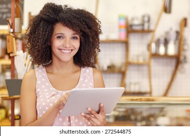Porträt einer schönen jungen Frau mit einer afro-Frisur, die bei der Eingabe ihres digitalen Tablets in ihrem Café die Kamera anlächelt