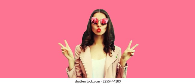 Portrait of beautiful young woman blowing her lips sending air kiss posing wearing sunglasses, leather jacket on pink background - Shutterstock ID 2255444183