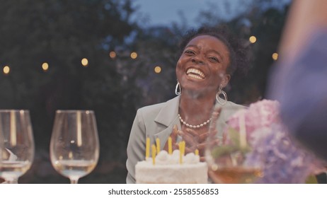 Portrait of beautiful young woman blowing candles on birthday cake. Happy African American girl celebrating her birthday with friends at home yard outdoors. Birthday party. Friends enjoying together. - Powered by Shutterstock