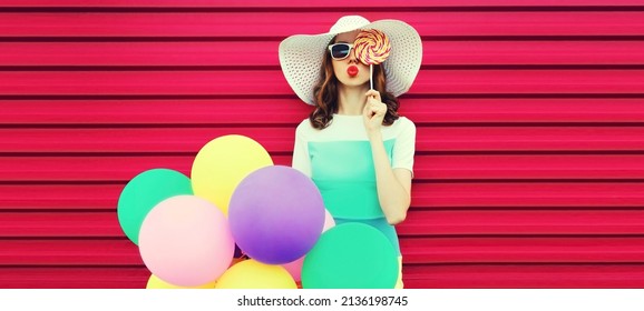 Portrait of beautiful young woman with bunch of balloons blowing her lips sends air kiss wearing a colorful dress on pink background