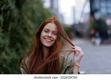 Portrait of a beautiful young woman in the city looking into the camera smile with teeth with red flying hair in fashionable clothes in the city against of bamboo in spring, lifestyle in the city