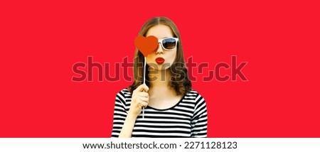 Portrait of beautiful young woman covering her eyes with red sweet heart shaped lollipop and blowing her lips on background
