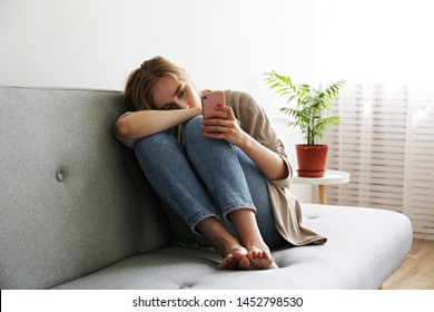 Porträt einer schönen jungen Frau mit depressiver Gesichtsausdruck sitzend auf grauem Textilcouch, die ihr Telefon hält. Cyber-Opfer-Konzept. Traurige Frau in ihrem Zimmer. Hintergrund, Kopienraum.