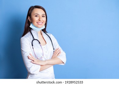 Portrait Of A Beautiful Young Woman Doctor In A White Robe On A Blue Background. A Place For Text And Advertising. Doctor Online. Medicine. Therapy And Pediatrics. High Quality Photo