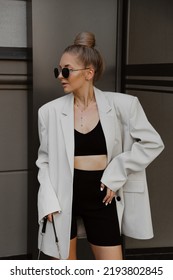 portrait of beautiful young woman dressed in beige oversized jacket, black knitted suit with shorts and top, bag, accessories, sunglasses, bun hairstyle, stylish summer fashion outfit, lifestyle model - Shutterstock ID 2193802845