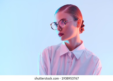 Portrait of a beautiful young woman in elegant glasses and white shirt on light blue background in pink lighting. Copy space. Business style. Glasses fashion. 