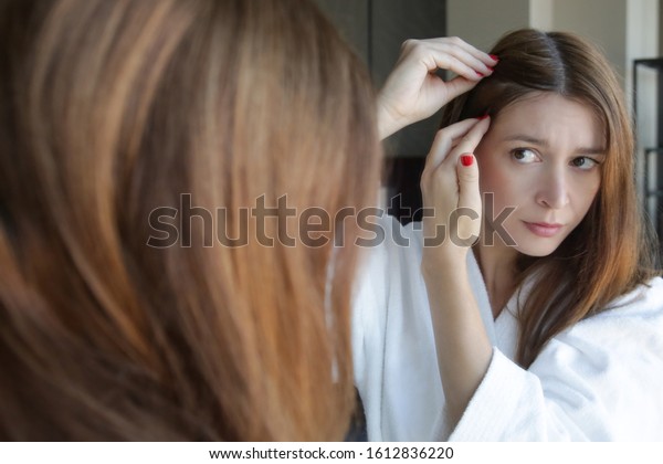 Portrait of a beautiful young woman examining her\
scalp and hair in front of the mirror, hair roots, color, grey\
hair, hair loss or dry scalp\
problem