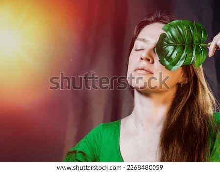 portrait of beautiful young woman eyes closed and monstera leaf on a half of the face, attractive brunette with long hair in a green dress. skin care beauty product concept