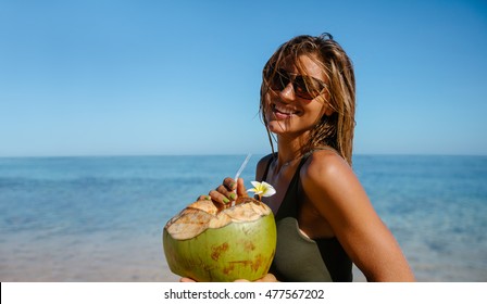 Portrait of beautiful young woman with fresh coconut on the beach. Caucasian woman wearing sunglasses having fresh coconut water on the sea shore.
