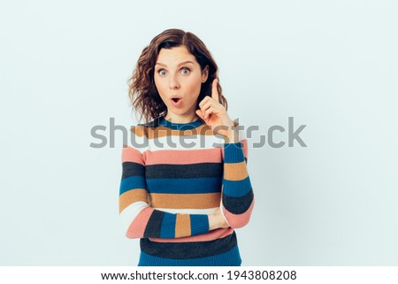 Portrait of beautiful young woman having an idea pointing with her index finger upwards with surprised expression on her face standing against white background.