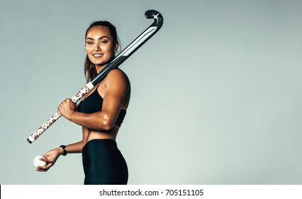 Portrait of beautiful young woman holding hockey stick and ball against grey background. Hispanic female hockey player looking at camera.