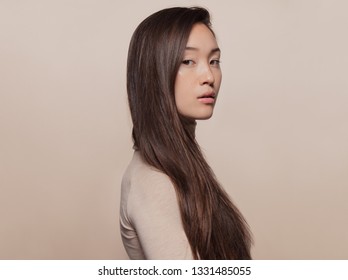 Portrait of beautiful young woman with long brown hair standing against beige background. Asian woman with a long straight hair looking at camera.