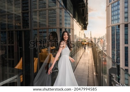 Portrait of beautiful young woman in long white dress standing on rooftop of building with cityscape scene