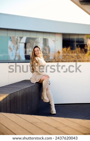 Portrait of a beautiful young woman with long wavy hair, wearing a short skirt and white blouse, posing on the terrace of a modern hotel