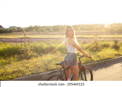 Portrait Of Beautiful Young Woman Looking Back Sitting On Mountain Bike Wearing Casual White Tank Top And Jeans Shorts On Countryside Road On Bright Sunny Summer Day