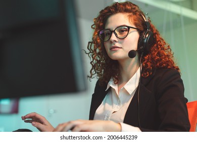 Portrait Of A Beautiful Young Woman Operator With Red Curly Hair And Glasses In The Interior Of The Call Center Office Works At The Computer And Uses A Special Headset For Calls