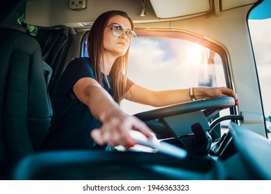 Portrait of beautiful young woman professional truck driver sitting and driving big truck. She is dangerously trying to take smart phone while driving.