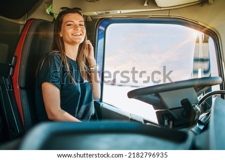 Portrait of beautiful young woman professional truck driver sitting in a big truck, looking at camera and smiling. Inside of vehicle. People and transportation concept.