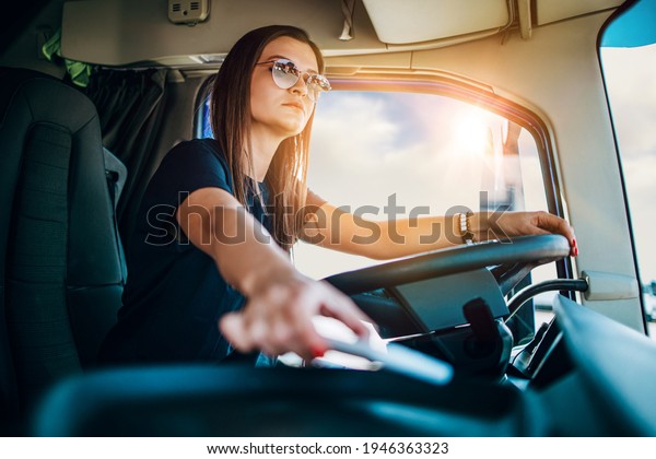 Portrait of beautiful young woman\
professional truck driver sitting and driving big truck. She is\
dangerously trying to take smart phone while\
driving.
