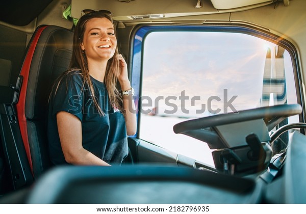 Portrait of beautiful young woman\
professional truck driver sitting in a big truck, looking at camera\
and smiling. Inside of vehicle. People and transportation\
concept.