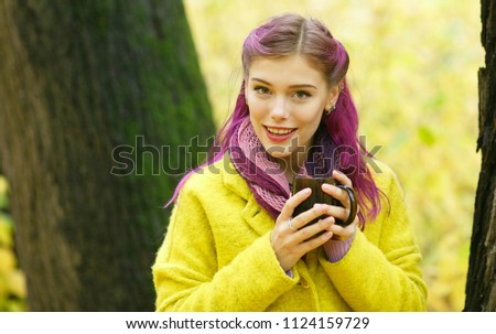 Portrait of a beautiful young woman with purple hair dressed in a yellow coat, looking with a smile at the camera, holding a mug with a hot drink while walking in the autumn forest. Autumn colors
