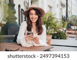 Portrait of beautiful young woman reading text message on mobile phone. Model at coffee shop. Female holds smartphone, uses apps. Outdoors in sunny day. In white blouse and hat. Cheerful and happy