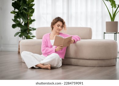 Portrait Of Beautiful Young Woman Reading Book While Relaxing At Home, Calm Millennial Female Sitting On Floor In Living Room And Enjoying Favorite Novel. Leisure, Literature And People Concept