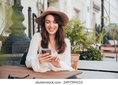 Portrait of beautiful young woman reading text message on mobile phone. Model at coffee shop. Female holds smartphone, uses apps. Outdoors in sunny day. In white blouse and hat. Cheerful and happy - Powered by Shutterstock