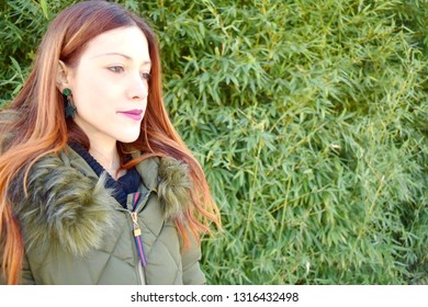 Portrait Of A Beautiful Young Woman With Red Hair And Dressing A Coat And With A Green Plant And Many Leaves On Background. Copy Space And Hdr Effect.