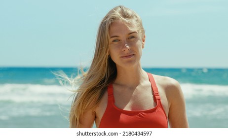 Portrait Of Beautiful Young Woman In Red Swimsuit Stand On Empty Sunny Beach, Look Into Camera With Confidence. Baywatch Or Coast Guard Tower In Background. Summertime Concept. Warm Breeze In Hair