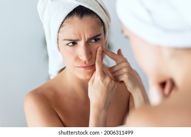 Portrait of beautiful young woman removing pimple from her face while looking at the mirror in a bathroom home.