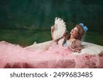 Portrait of beautiful young woman, royal person, princess in elegant dress lying on bed with feather fan and with flirty face, against vintage green background. Concept of history, comparison of eras