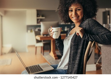 Portrait of beautiful young woman sitting at home with laptop and having coffee. African female relaxing at home with coffee and laptop looking at camera and smiling. - Shutterstock ID 1277145427